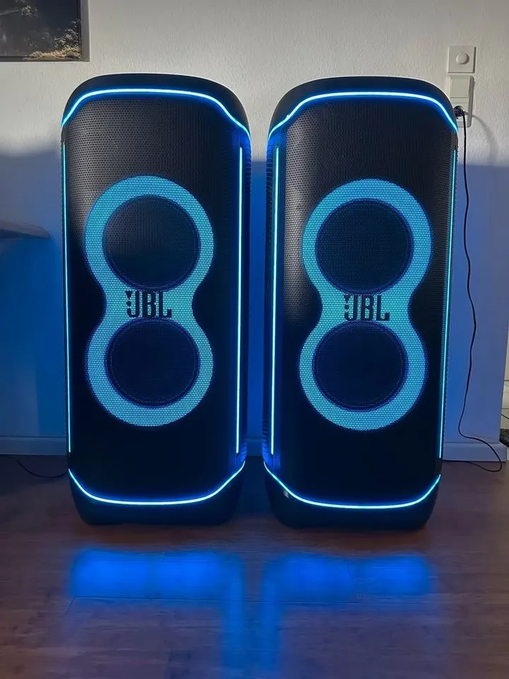 Zwei JBL PartyBoxen nebeneinander in einem Wohnzimmer, die Ringe leuchten türkis