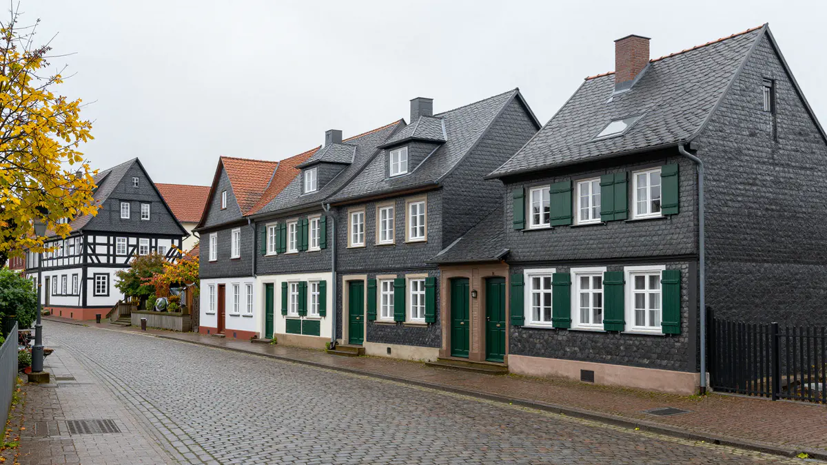 Illustration: Gepflasterte Straße mit Schieferhäusern, grünen Fensterläden und einem Fachwerkhaus an einem trüben Herbsttag
