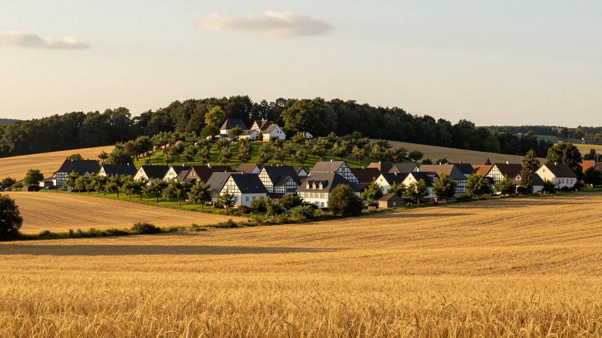 Illustration: Dorf mit Fachwerkgiebeln und Obstwiese auf einer Anhöhe hinter goldenen Getreidefeldern im Abendlicht
