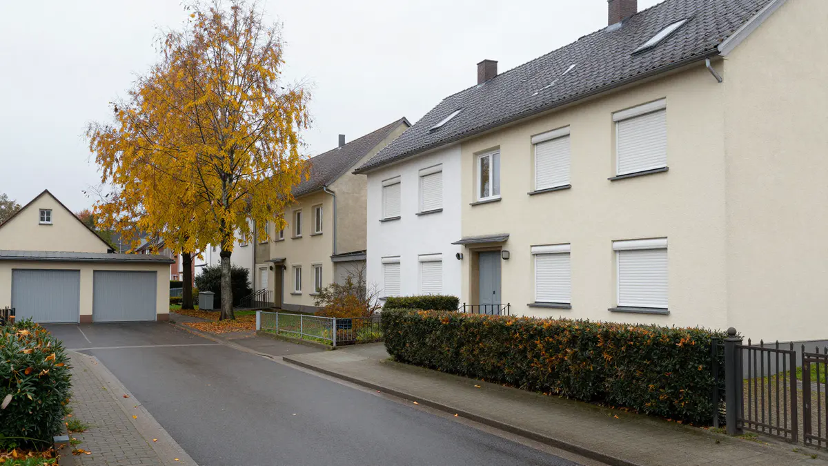 Illustration: Ruhige Wohnstraße mit hell verputzten Wohnhäusern, Rollläden, Garagenhof und gelb belaubtem Baum im Herbst