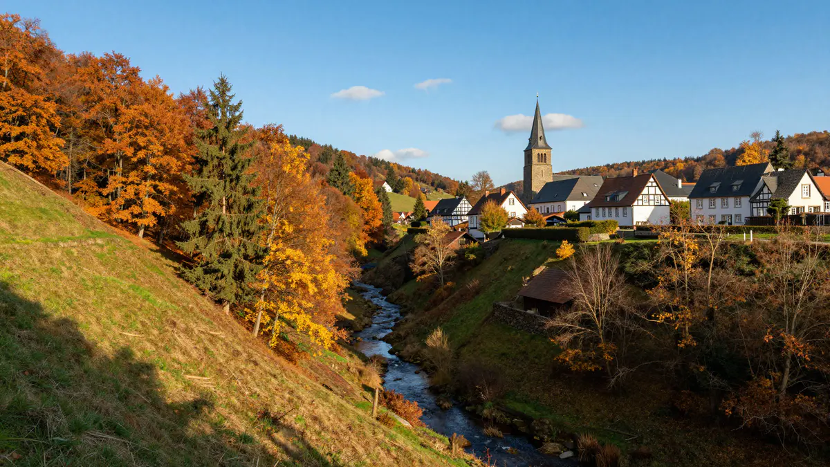 Illustration: Blick in ein Bachtal mit Herbstwald, auf der Anhöhe gegenüber ein Kirchdorf mit Fachwerkhäusern und spitzem Kirchturm
