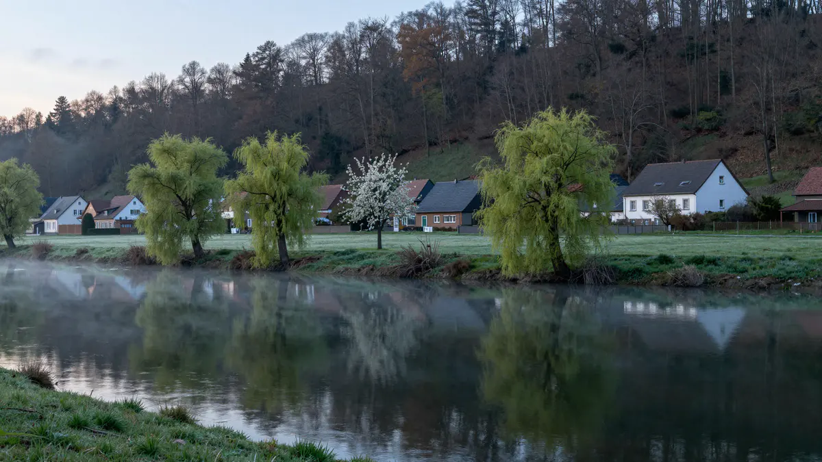 Illustration: Ruhiger Fluss im Morgendunst mit Weiden am Ufer, blühendem Obstbaum und Wohnhäusern vor bewaldetem Hang