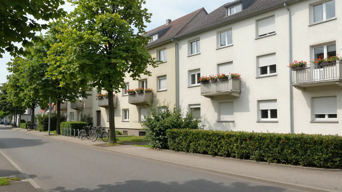 Illustration: Ruhige Wohnstraße mit hellen Mehrfamilienhäusern, Balkonen mit Geranien, Straßenbäumen und Fahrrädern