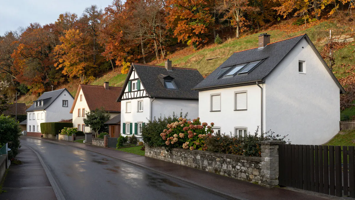 Illustration: Regennasse Wohnstraße mit weißen Häusern, Schieferdächern und einem Fachwerkgiebel vor herbstlichem Hangwald