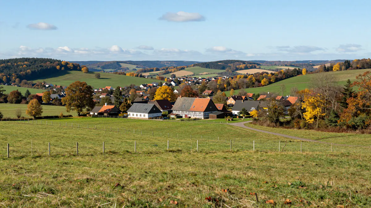 Illustration: Weiter Blick über Weiden mit Zaunpfählen auf ein Dorf mit Fachwerkscheune und bewaldete Hügel im Herbst