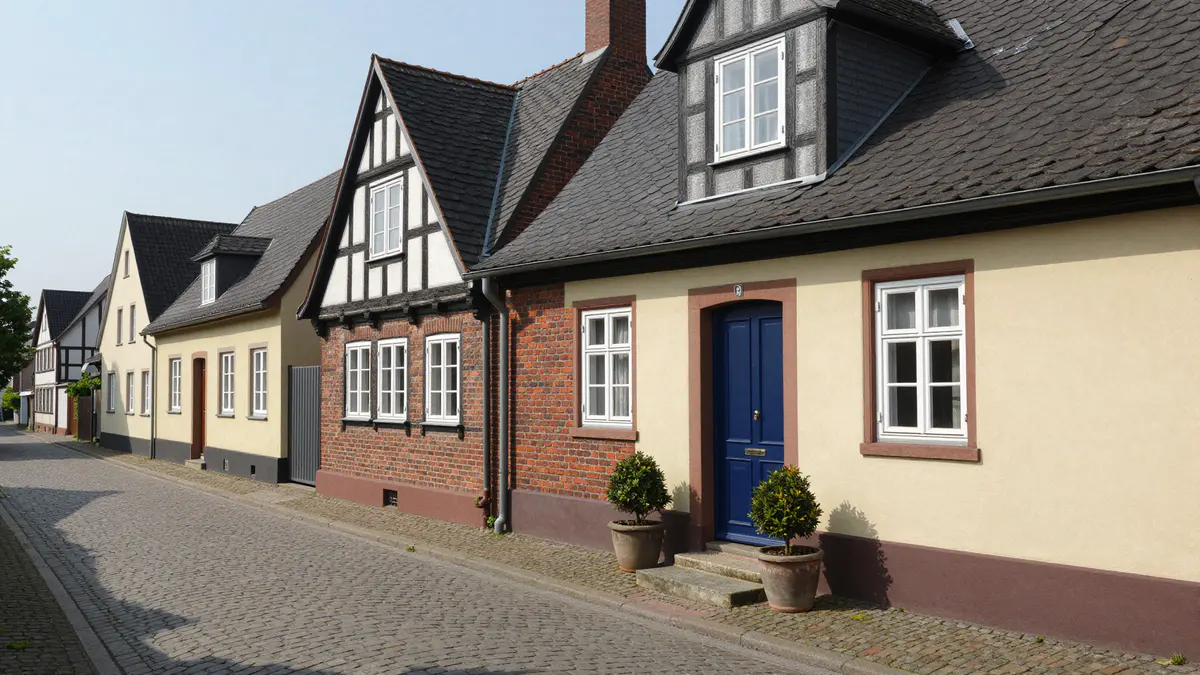Illustration: Gepflasterte Altstadtgasse mit hellen Putzhäusern, Backstein und Fachwerk, vor einer blauen Haustür zwei Buchsbäume in Töpfen