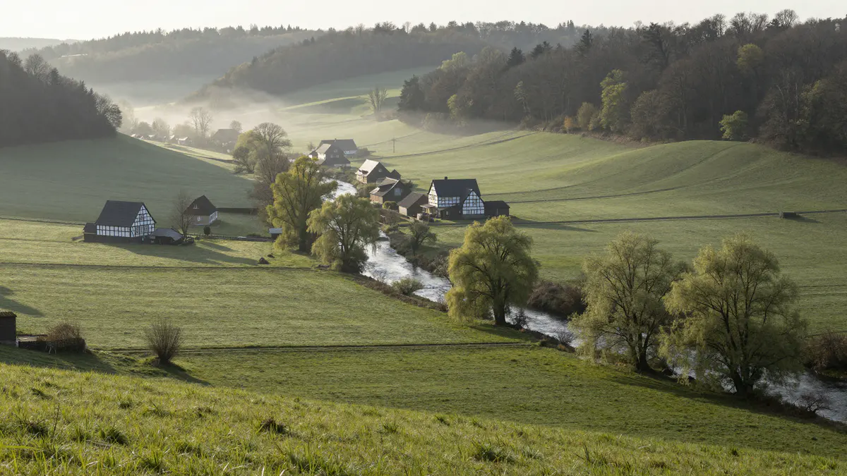 Illustration: Blick von einer Wiese ins Tal mit gewundenem Fluss, Weiden, Fachwerkhöfen und bewaldeten Höhen im Morgendunst