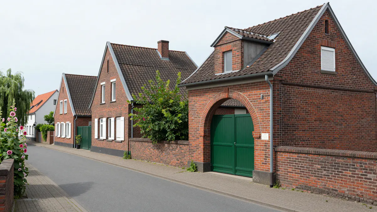 Illustration: Ruhige Dorfstraße mit roten Backsteinhäusern, weißen Fensterläden und einem Hof mit grünem Holztor im Rundbogen
