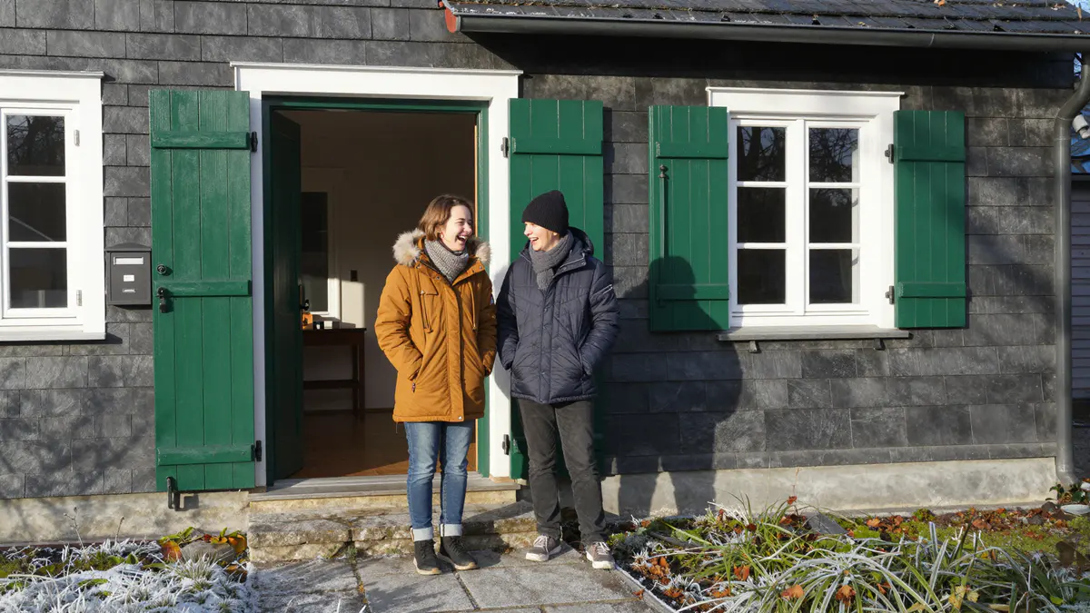 Illustration: Zwei lachende Menschen in Winterjacken stehen vor der offenen grünen Tür eines schieferverkleideten Hauses
