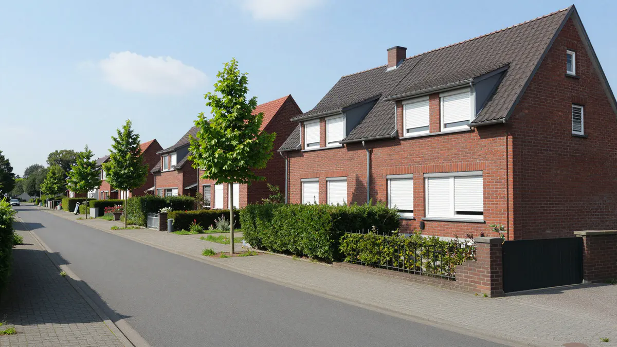 Illustration: Ruhige Wohnstraße mit roten Klinkerhäusern, weißen Rollläden, Hecken und jungen Straßenbäumen im Sommer